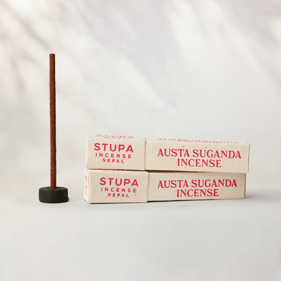 Two paper boxes of Stupa Austha Suganda incense beside a stick standing in a small holder, on a pale background.