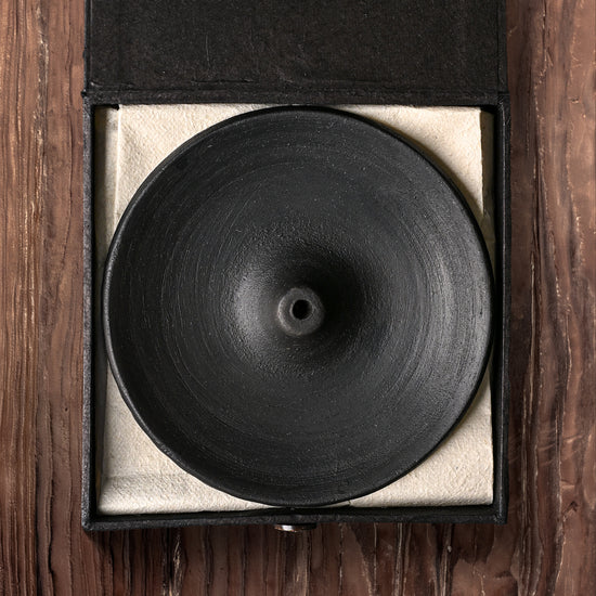 Clay incense stick holder: round black dish in its open black box with a pale lining, on a wooden surface.