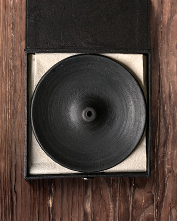 Clay incense stick holder: round black dish in its open black box with a pale lining, on a wooden surface.