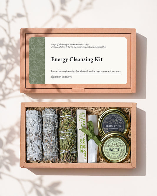An open wooden gift box labeled 'Energy Cleansing Kit' by Morning Visions, containing a sage bundle, a dried herb bundle, a white crystal candle, a green sprig, and two round tins, arranged on a light neutral background with soft leaf shadows.