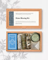 Home Blessing Kit: kraft box lid above the open box with sandalwood incense, three smudge sticks, a frankincense tin and a white crystal pyramid.