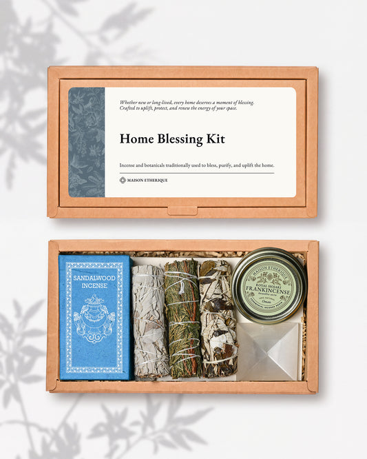 An open wooden gift box labeled 'Home Blessing Kit' containing a blue incense box, sage bundles, a round tin, and a white crystal, photographed on a light gray background with soft shadow overlays.
