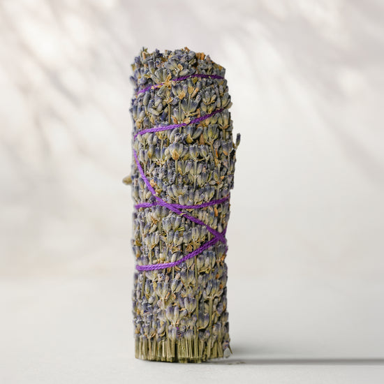 A lavender smudge bundle wrapped with purple string, standing upright on a light gray surface against a soft, shadowed light gray background.