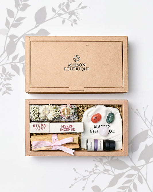 An open Maison Etherique gift box containing a crystal cluster, a white sage with sachet of dried flowers, and lavender essential oil  arranged in a kraft paper tray with the matching lid resting above on a light background with faint leafy illustrations.