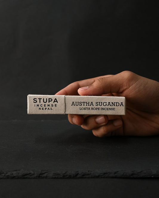 Hand holding a box of Stupa Austha Suganda lokta rope incense against a dark background.