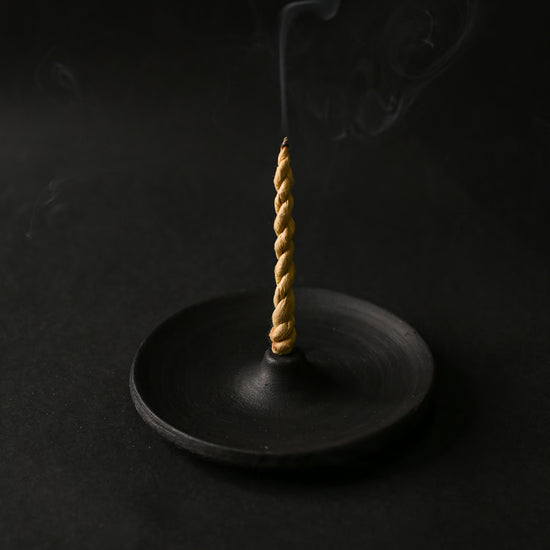 Twisted rope incense burning upright in a round black clay incense holder, smoke rising against a dark background.