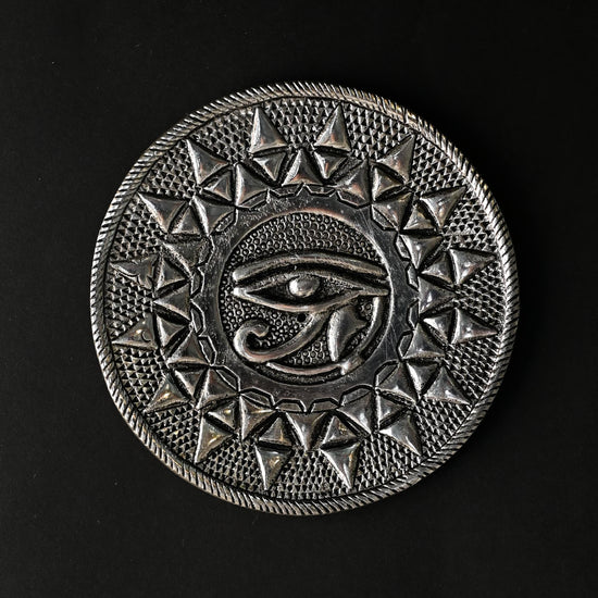 Eye of Horus incense stick holder, a round silver-toned metal plate with an embossed eye at its centre, on a dark background.
