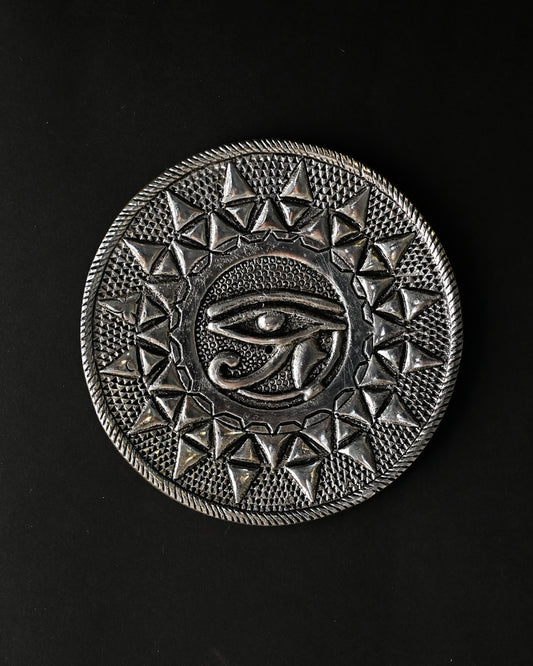 Eye of Horus incense stick holder, a round silver-toned metal plate with an embossed eye at its centre, on a dark background.