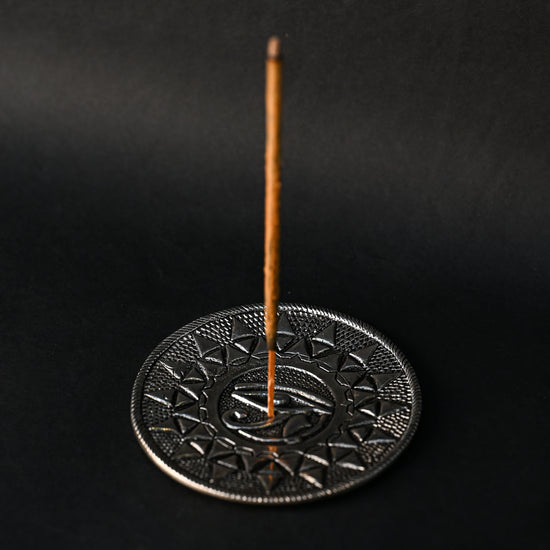 Incense stick standing upright in the Eye of Horus metal holder against a dark background.
