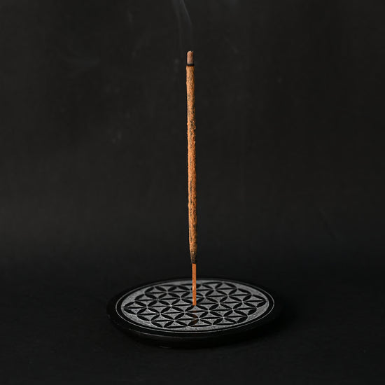 Incense stick burning upright in the round Flower of Life stone holder, smoke rising against a dark background.