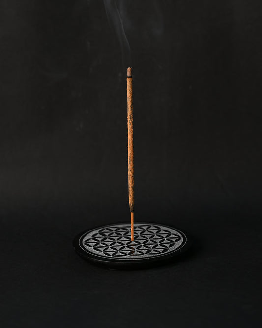 Incense stick burning upright in the round Flower of Life stone holder, smoke rising against a dark background.