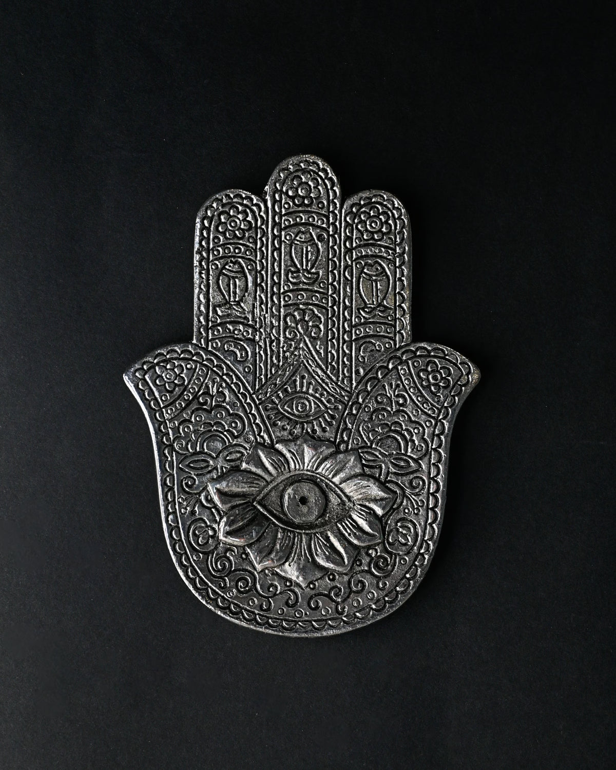 Hand-shaped incense stick holder in silver-toned metal with an eye motif at its centre, seen from above on a dark background.