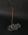 Incense stick burning upright in the hand-shaped metal incense holder, smoke rising against a dark background.