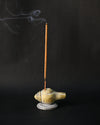 Incense stick burning upright in the conch-shaped Shankh stone holder, smoke drifting against a dark background.