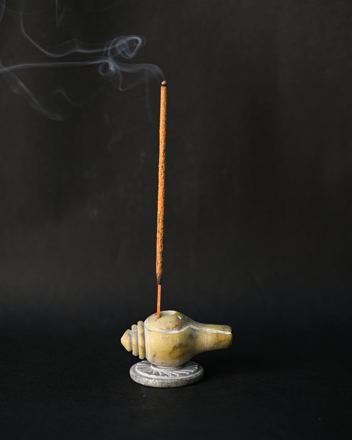 Incense stick burning upright in the conch-shaped Shankh stone holder, smoke drifting against a dark background.
