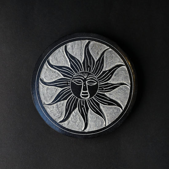 Soleil Noir incense stick holder, a round black stone disc carved with a sun face, seen from above on a dark background.