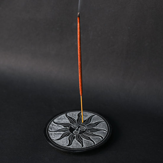 Incense stick burning upright in the Soleil Noir carved stone holder, smoke rising against a dark background.