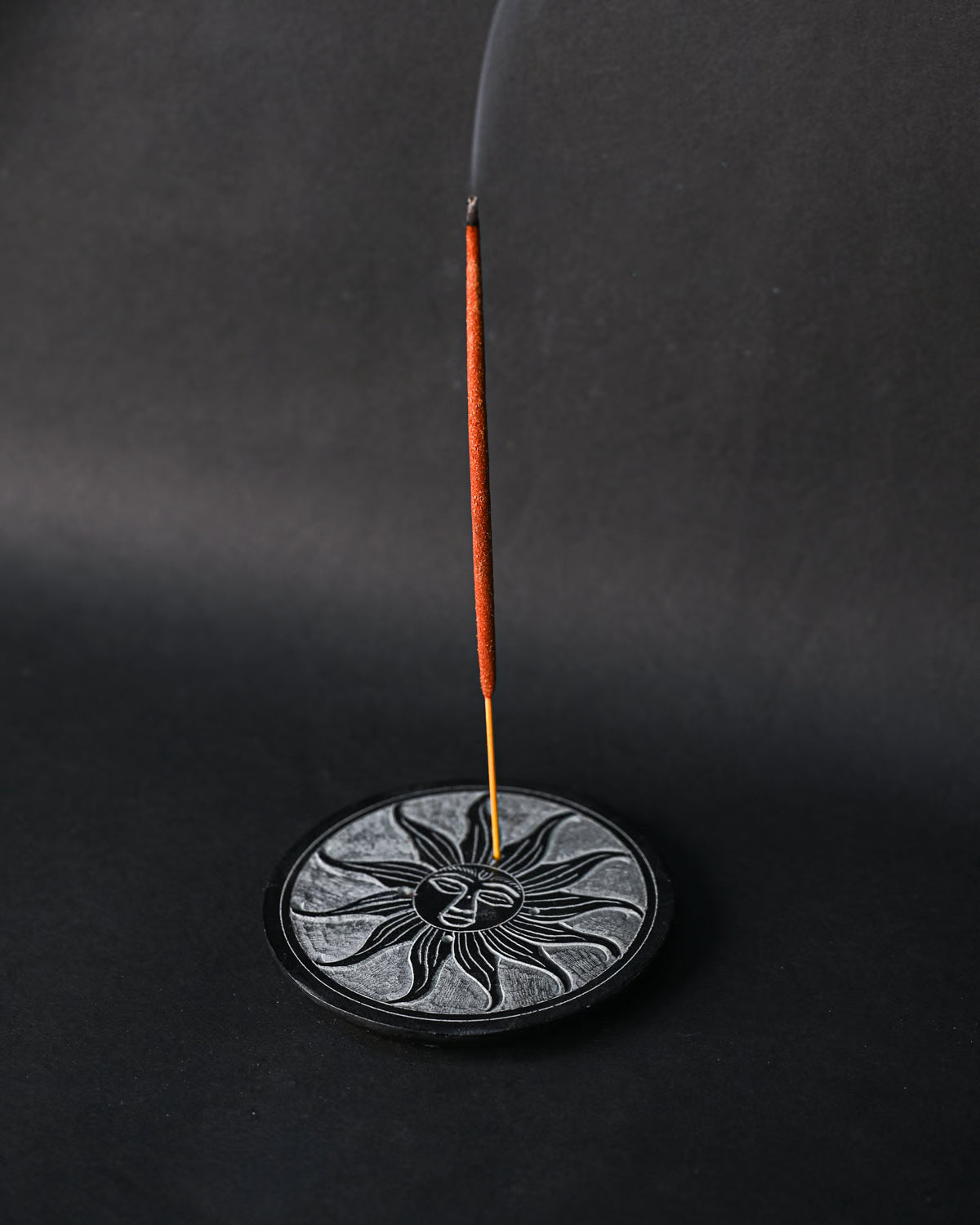 Incense stick burning upright in the Soleil Noir carved stone holder, smoke rising against a dark background.