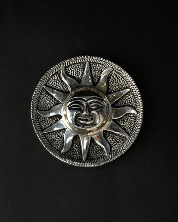 Surya incense stick holder, a round silver-toned metal plate with a sun face at its centre, on a dark background.