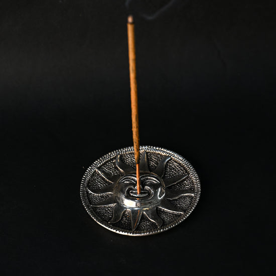 Incense stick burning upright in the Surya sun-face metal holder, smoke rising against a dark background.