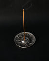Incense stick burning upright in the Surya sun-face metal holder, smoke rising against a dark background.