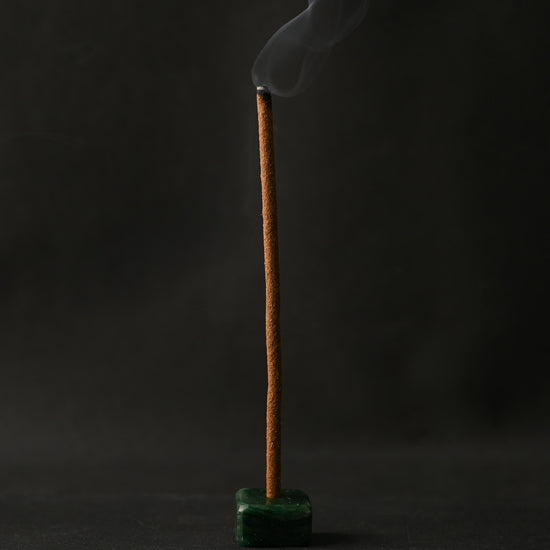 Padma Tibetan incense stick burning upright in a small green holder, smoke curling against a dark background.