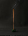 Padma Tibetan incense stick burning upright in a small green holder, smoke curling against a dark background.