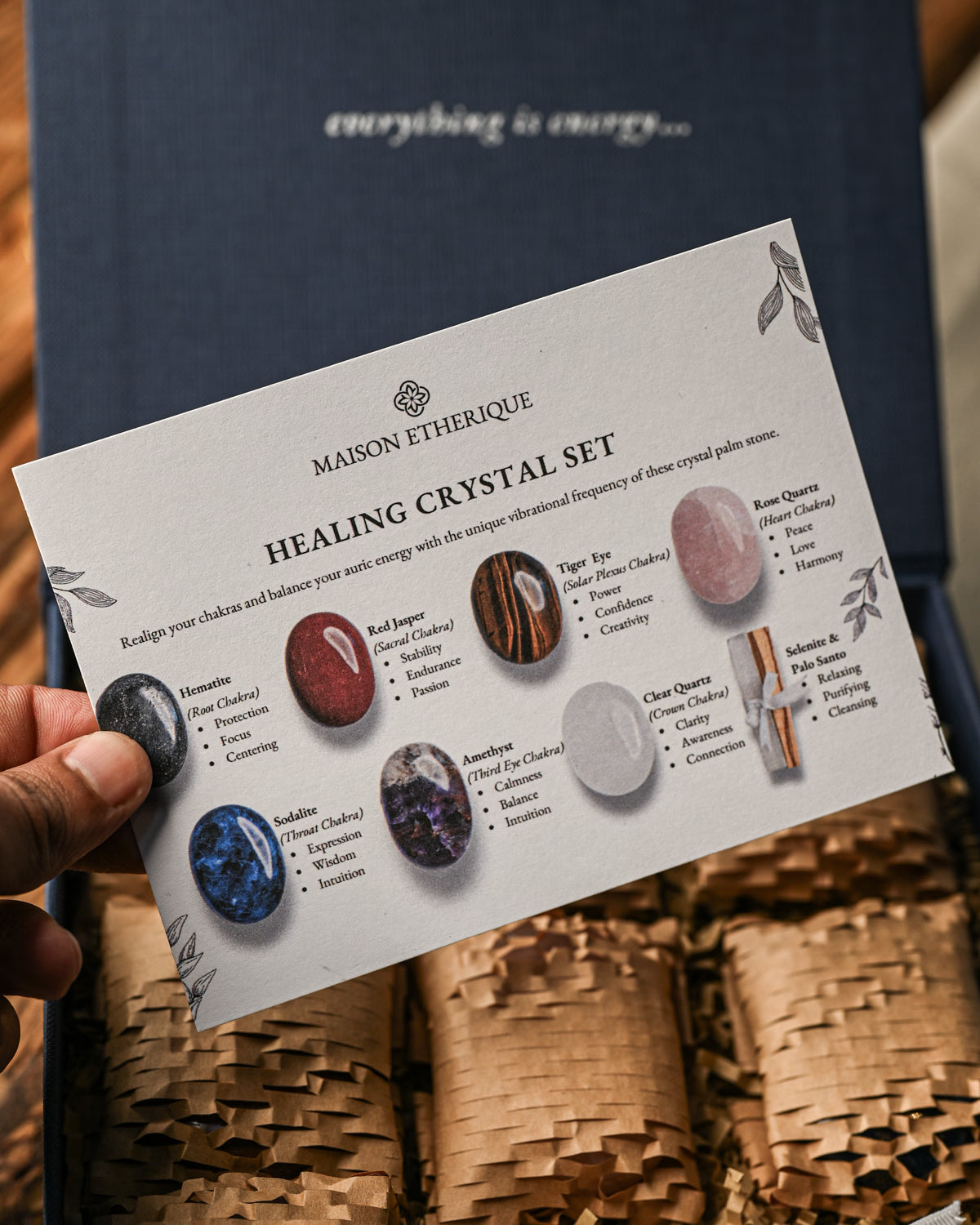 Hand holding the printed guide card of the Maison Éthérique palm stones crystal set above its paper-lined box.