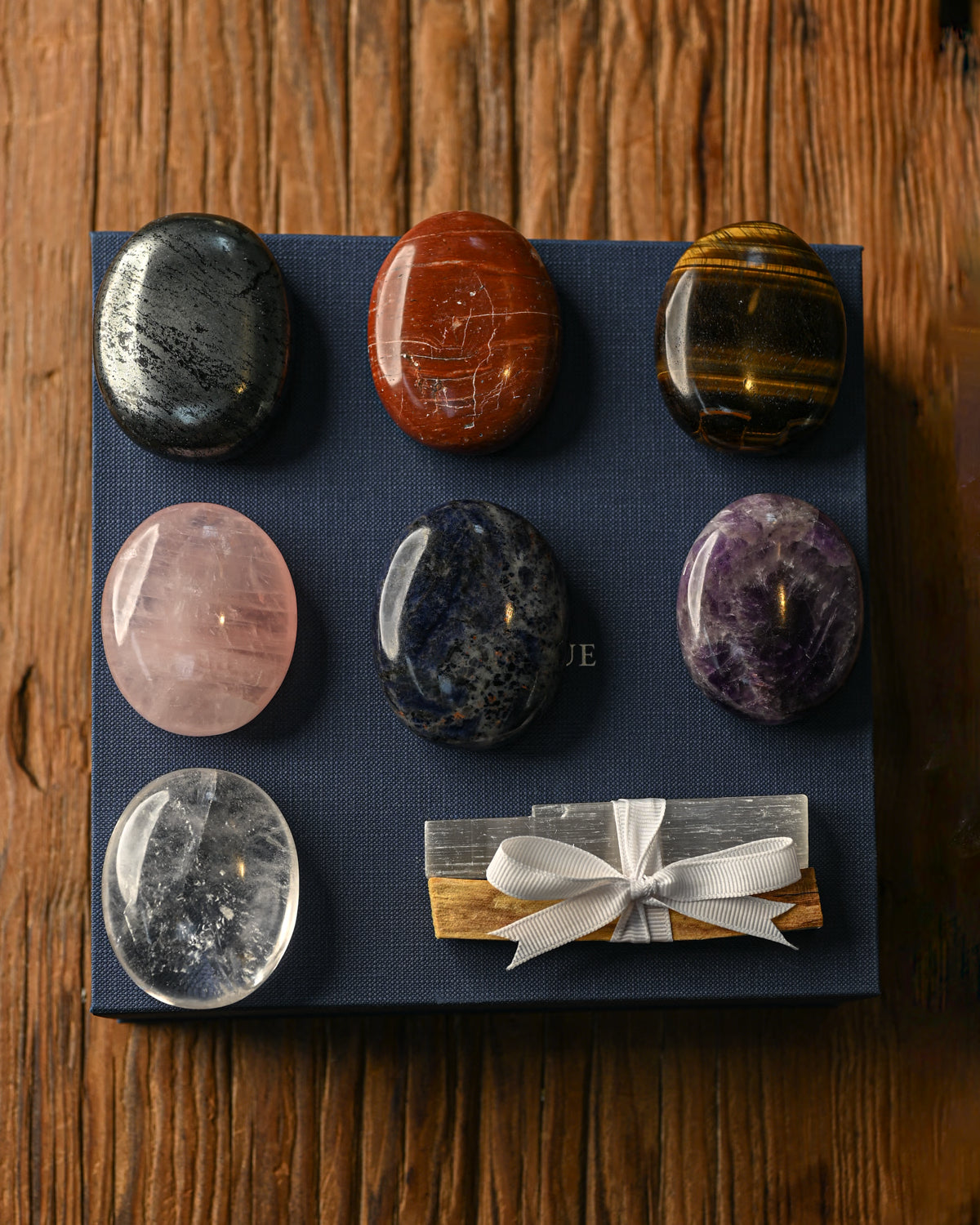 Seven polished palm stones and a ribbon-tied Palo Santo bundle arranged on a navy box, on a wooden surface.