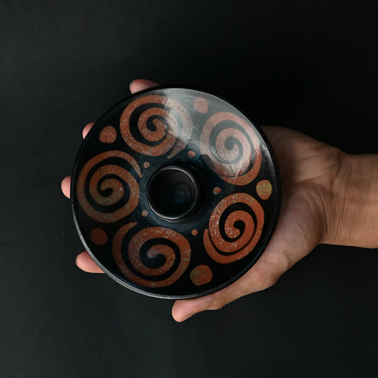 Hand holding a black ceramic Palo Santo burner painted with rust-red spirals against a dark background.