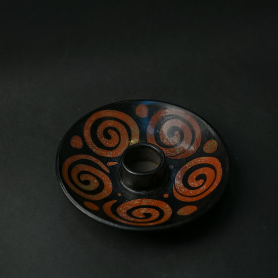 Black ceramic Palo Santo burner painted with rust-red spirals, seen at an angle on a dark background.
