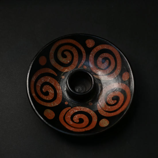 Black ceramic Palo Santo burner painted with rust-red spirals, seen from above on a dark background.