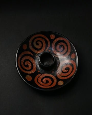 Black ceramic Palo Santo burner painted with rust-red spirals, seen from above on a dark background.