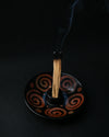 Palo Santo stick smouldering upright in a black ceramic burner painted with rust-red spirals, smoke rising.