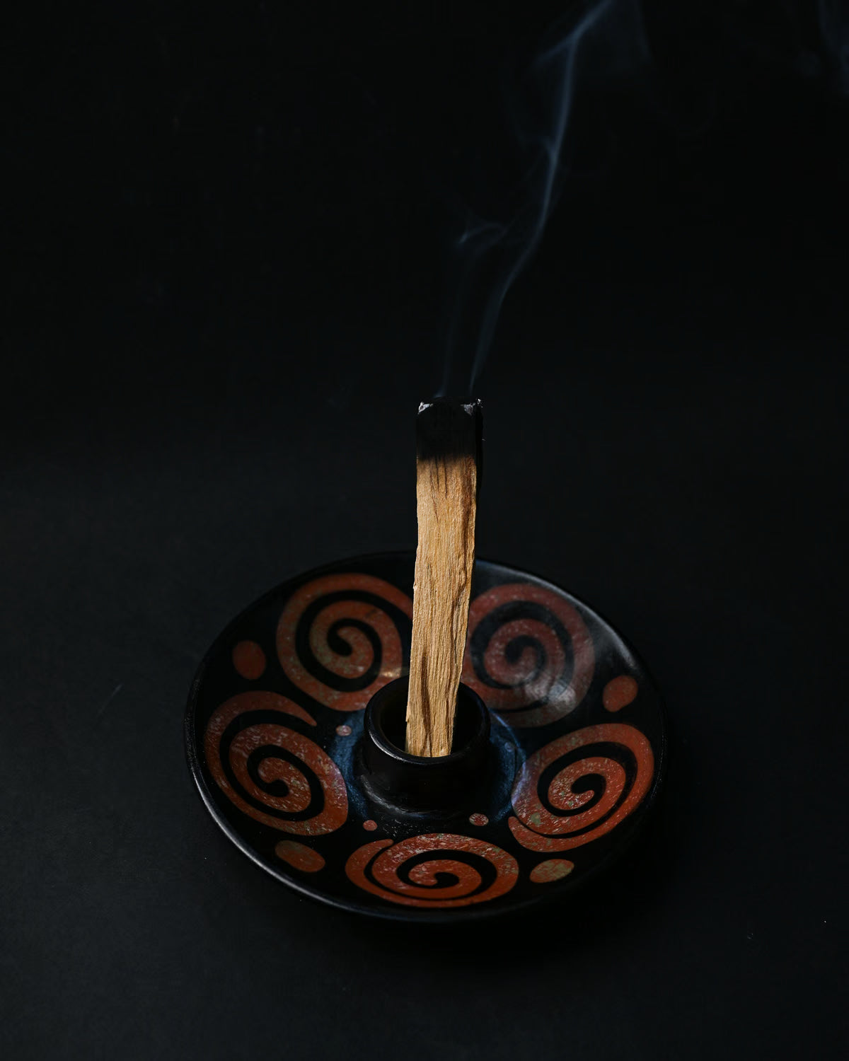 Palo Santo stick smouldering upright in a black ceramic burner painted with rust-red spirals, smoke rising.