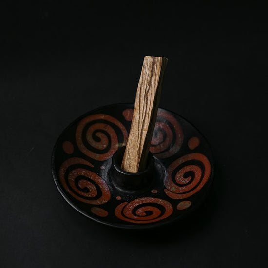 Palo Santo stick standing in a black ceramic burner painted with rust-red spirals, on a dark background.