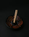 Palo Santo stick standing in a black ceramic burner painted with rust-red spirals, on a dark background.