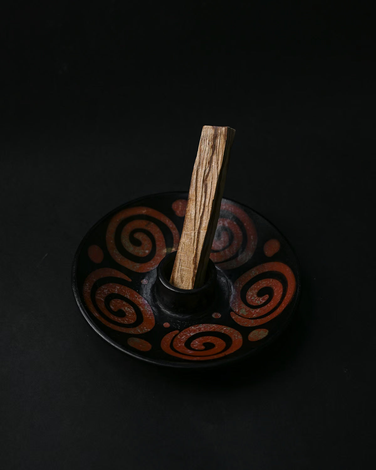 Palo Santo stick standing in a black ceramic burner painted with rust-red spirals, on a dark background.