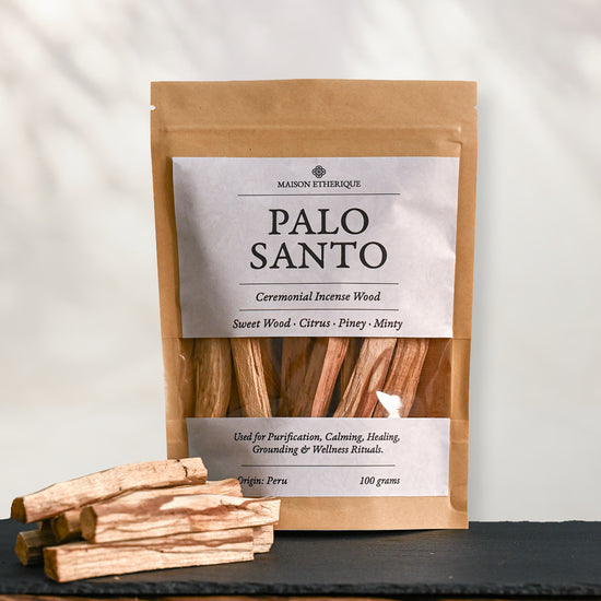 Kraft pouch of Maison Éthérique Palo Santo, 100 g, standing on dark slate with loose Palo Santo sticks in front.