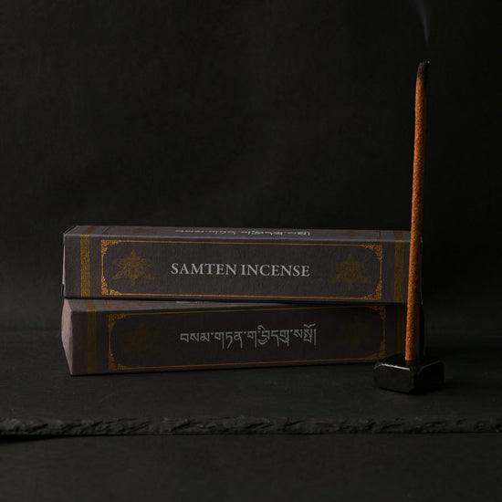 Two dark boxes of Samten Tibetan incense beside a stick burning upright in a small holder, on a dark background.
