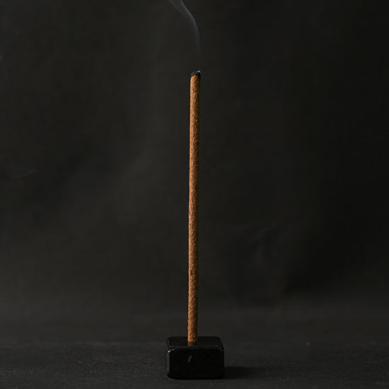 Samten Tibetan incense stick burning upright in a small dark holder, smoke rising against a dark background.