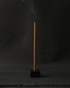 Samten Tibetan incense stick burning upright in a small dark holder, smoke rising against a dark background.