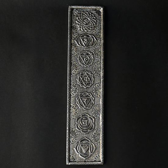 7 Chakra incense stick holder in silver-toned metal, embossed with seven symbols, seen from above on a dark background.
