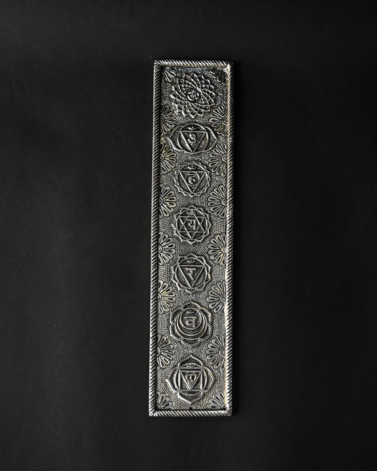 7 Chakra incense stick holder in silver-toned metal, embossed with seven symbols, seen from above on a dark background.