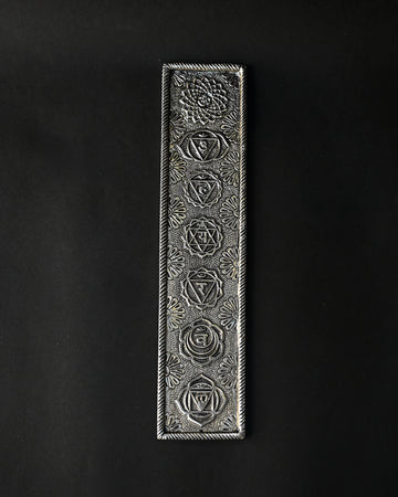 7 Chakra incense stick holder in silver-toned metal, embossed with seven symbols, seen from above on a dark background.